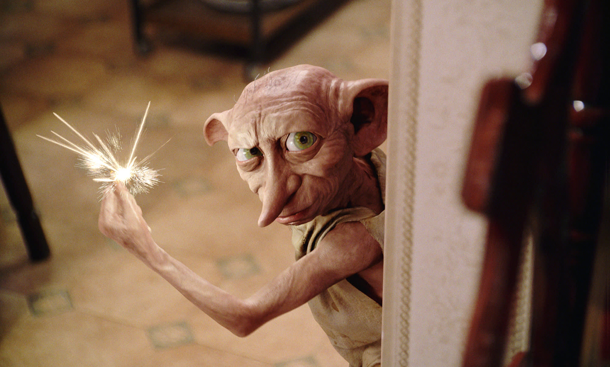 Dobby is standing in a doorway at Privet Drive. He is snapping his fingers while sparks fly from them.